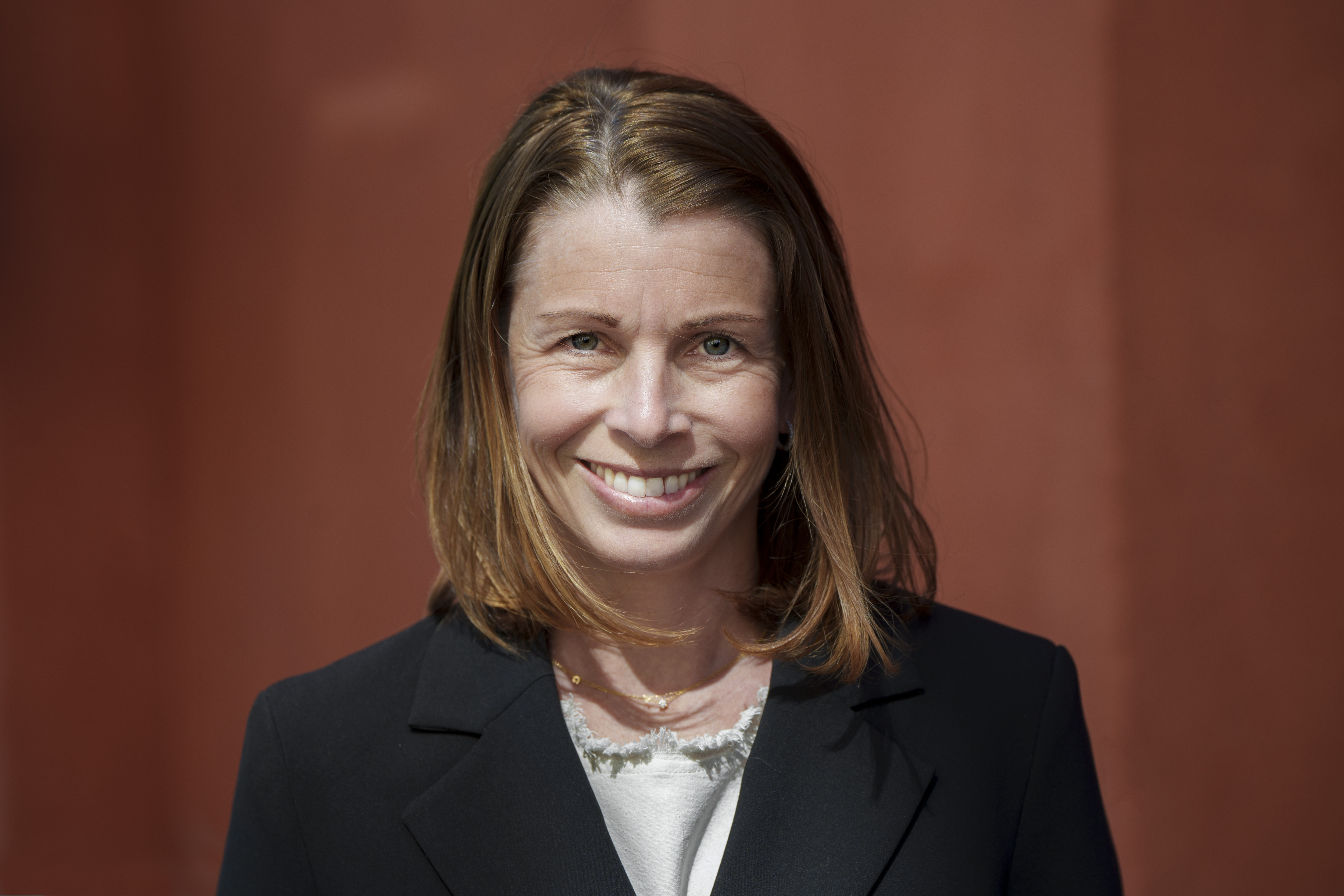 Närbild på en leende Malin Söderström framför en rödfärgad bakgrund. Hon har axellångt brunt hår och är klädd i mörk kavaj och en vit blus.